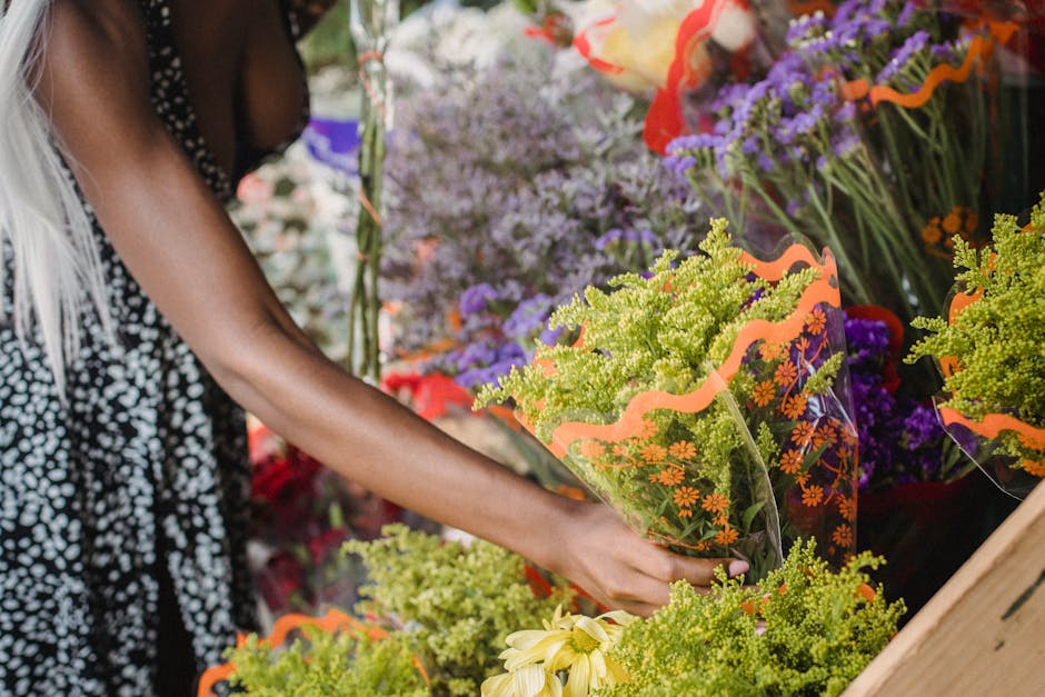 A person with dark skin and long, blonde hair selecting a bouquet of fresh flowers at a floral shop. The bouquet features vibrant green foliage and small orange flowers, wrapped in clear cellophane wi