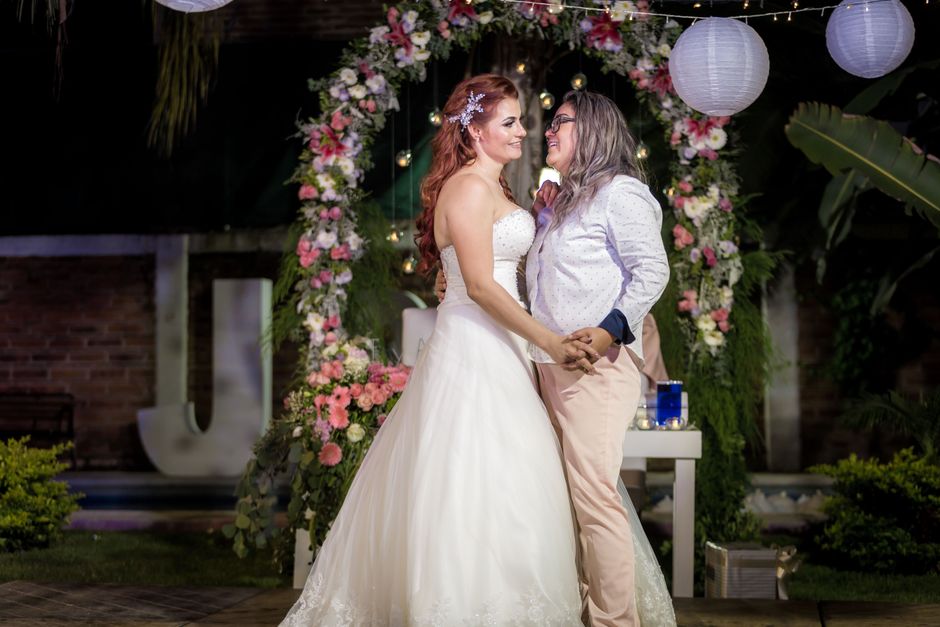 A bride with long, wavy red hair wearing a strapless white wedding gown and a delicate floral hairpiece is holding hands with a woman who has shoulder-length gray hair and glasses, dressed in a white 