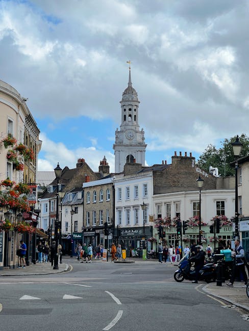 The image shows a lively street scene in a historic town with a prominent clock tower featuring a spire under a partly cloudy sky. Surrounding the tower are charming, traditional buildings with a mix 