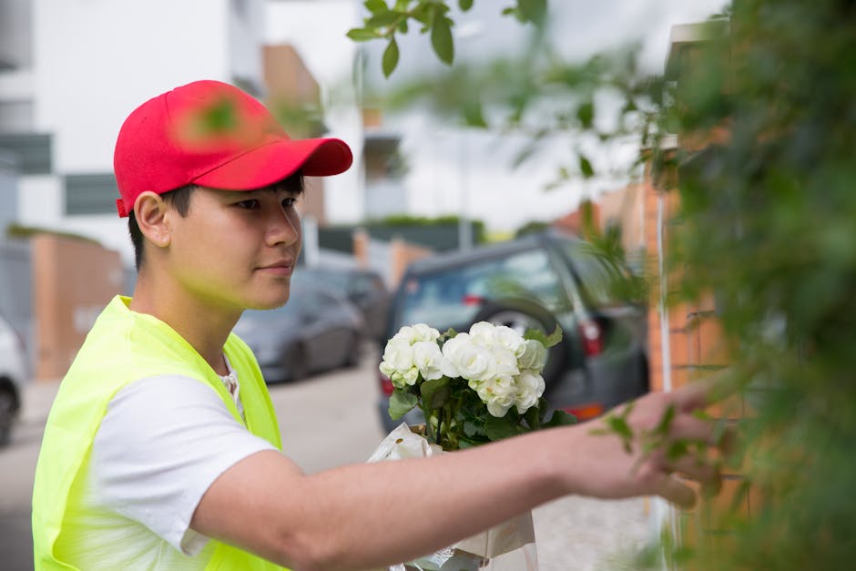 A young delivery person wearing a red cap and a high-visibility yellow vest is seen placing a bouquet of white roses and hydrangeas into a green bush near a residential street. The bouquet is wrapped