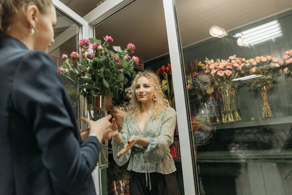 A florist with blonde curly hair, wearing a light-colored floral blouse, hand gestures while speaking to a customer, inside a flower shop with glass display cases filled with pink roses and other flor